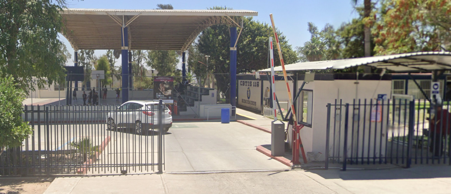 Fotografía en la que se muestra la entrada del plantel que se conforma por una reja color azul, y que a la mitad tiene un espacio que permite la entrada de los carros. Al fondo se pueden ver unas canchas techadas.